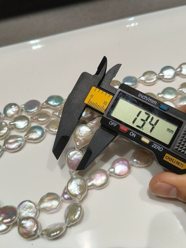 A digital caliper measuring a 13.4mm metallic coin baroque pearl with vibrant rainbow overtones on a strand.