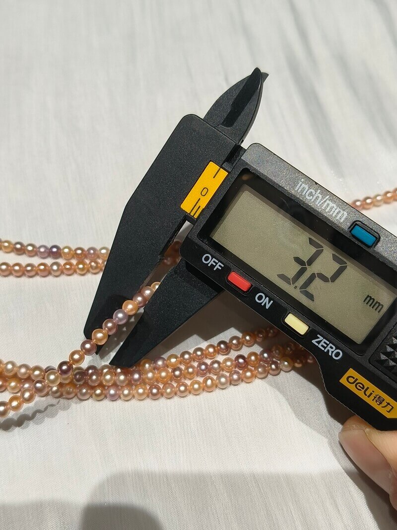 A digital caliper measuring the 3.2mm size of miniature multi-color freshwater seed pearls on a strand.